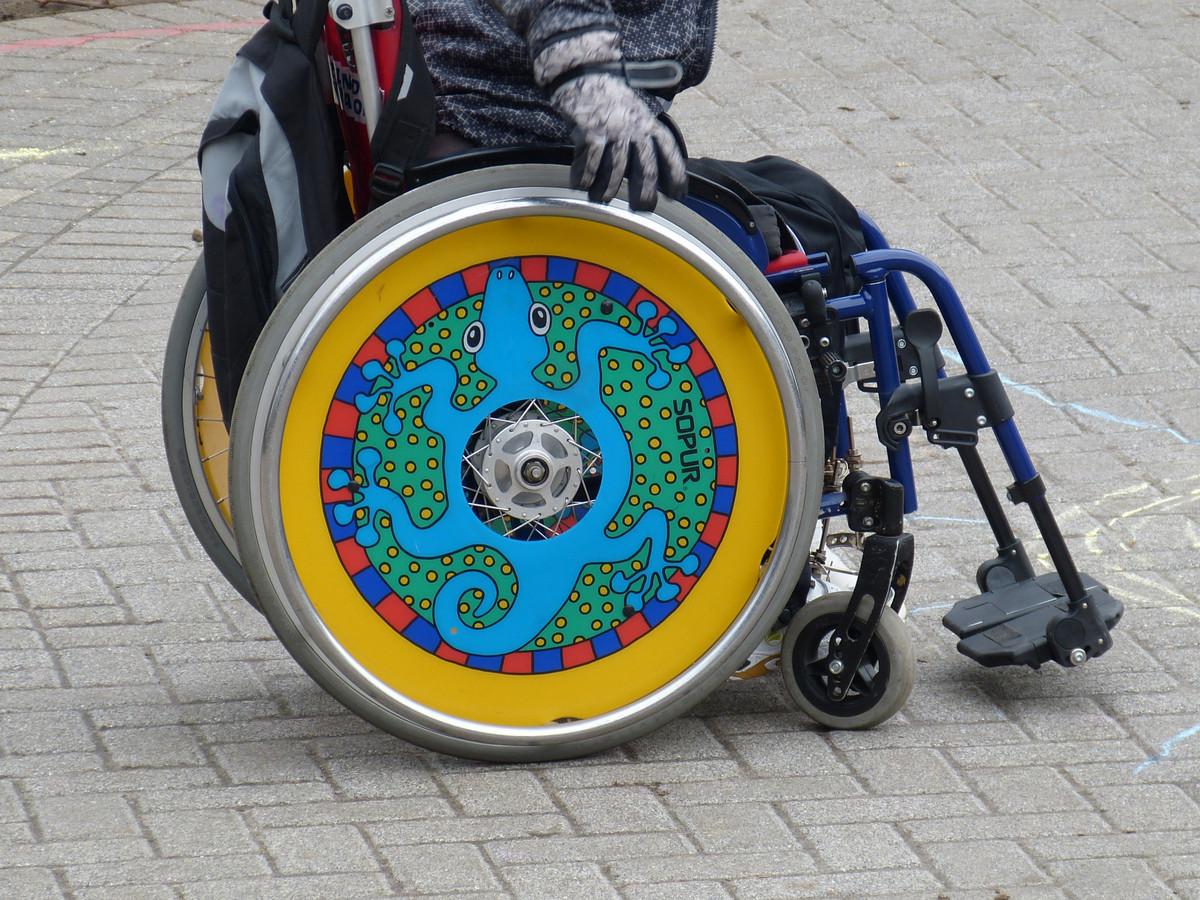 Ein Rollstuhl mit bunten Radscheiben von der Seite, im Rollstuhl sitzt eine Person, die man bis zum Oberkörper sehen kann.