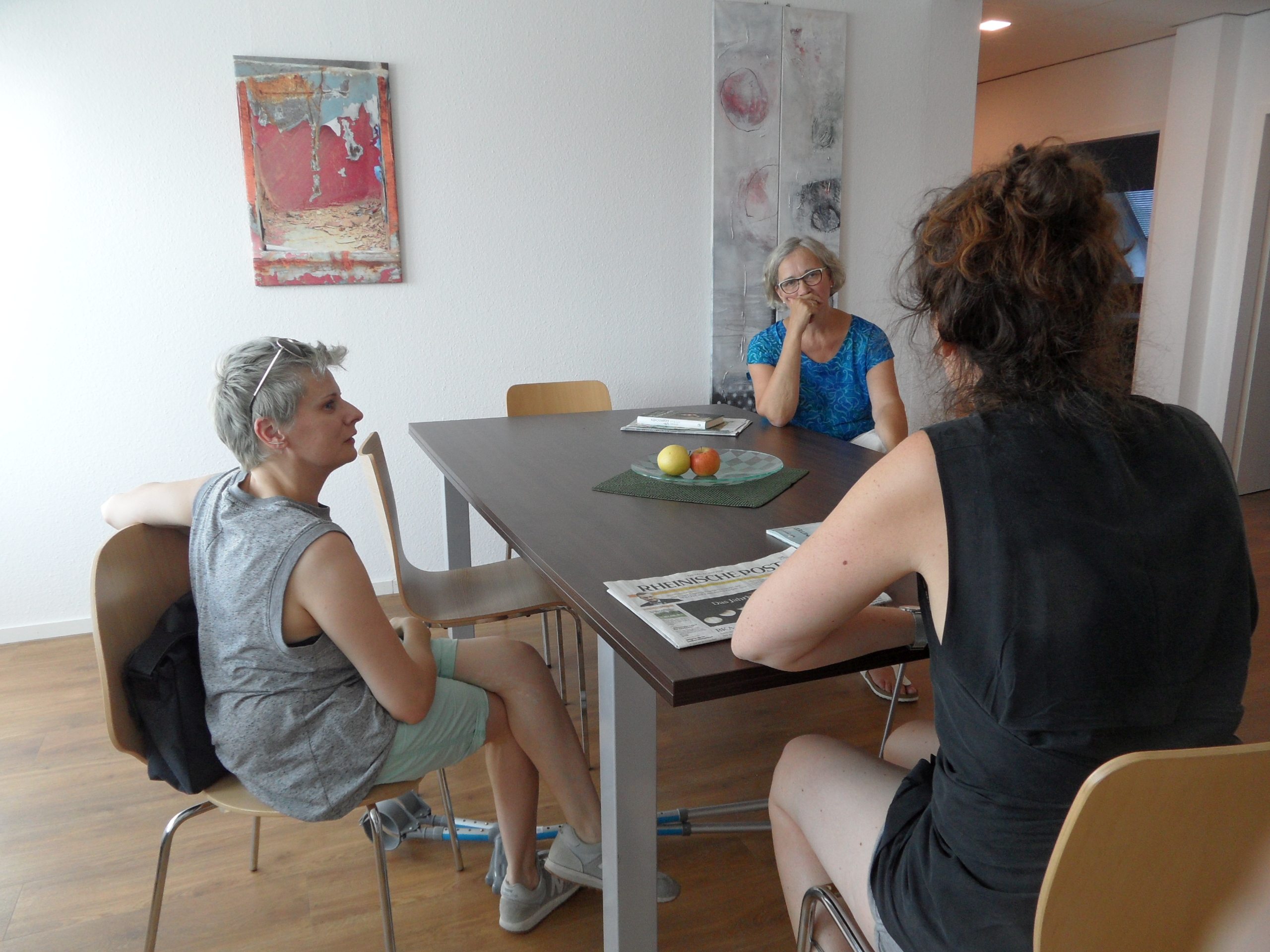 Links sitzt Claire Cunningham an einem Tisch, rechts am Tisch zwei Frauen, ein Gespräch