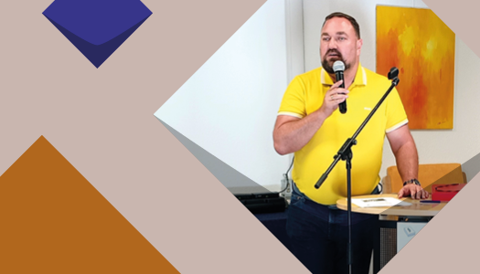 Eine braune Kachel der KSL und ein Foto von einem Mann mit kurzen brauen Haaren und einem gelben T-Shirt mit einem Microphone während eines Vortrages