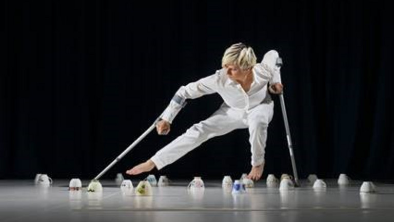 Die schottische Performance-Künstlerin Claire Cunningham im weißen Hosenanzug tanzt mit zwei Krücken zwischen umgedrehten Porzellantassen auf der Bühne - Foto: Colin Mearns