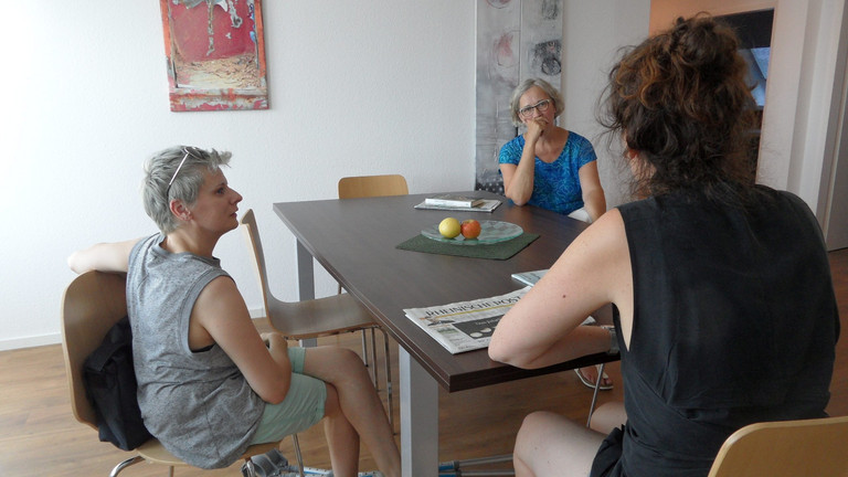 Links sitzt Claire Cunningham an einem Tisch, rechts am Tisch zwei Frauen, ein Gespräch