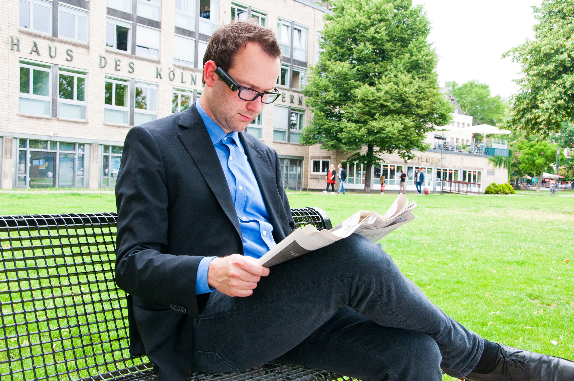 Ein Herr sitzt auf einer Bank und liest Zeitung. Er trägt eine intelligente Brille, eine Kamera ist am Bügel befestigt.