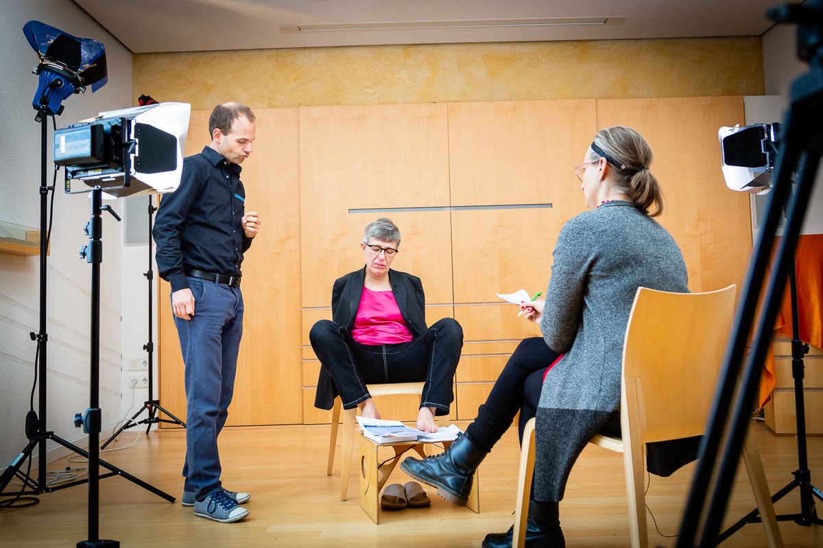 Eine Vorbereitung auf ein Interview. In der Mitte sitzt Frau Degener. Vor ihr sitzt Frau Roth, die Interviewerin. Links von Frau Degener steht ein Mann.