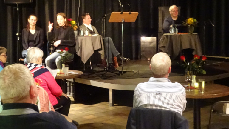 Das Bild zeigt Axel Brauns (rechts auf der Bühne), Olaf Reitz (links auf der Bühne), Iris Colsman, Leiterin des KSL Düsseldorf (rechts im Bild), zwei Gebärdensprachdolmetscherinnen (links im Bild) und einige Zuschauer*innen.