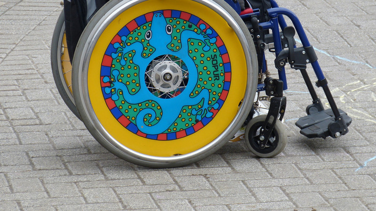 Ein Rollstuhl mit bunten Radscheiben von der Seite, im Rollstuhl sitzt eine Person, die man bis zum Oberkörper sehen kann.