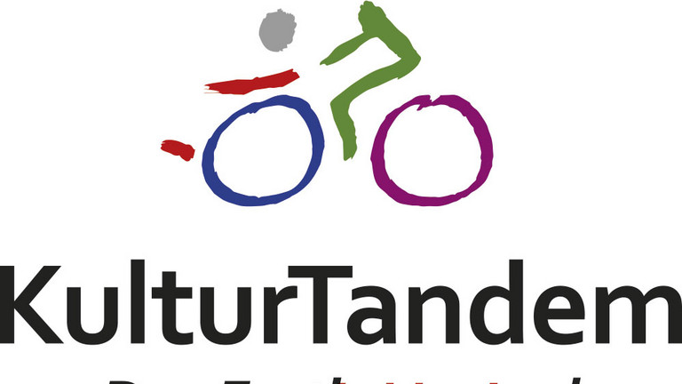 Logo KulturTandem: Ein stilisiertes Rollstuhlfahrrad mit wenigen Pinselstrichen gemalt, mit dem von rechts nach links zwei Personen fahren